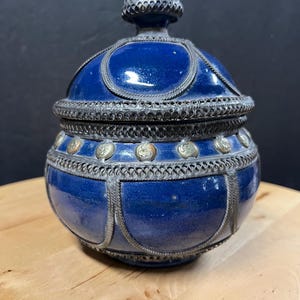 Può includere: Un vaso in ceramica blu cobalto con un design a filigrana in metallo argentato. Il vaso ha una forma arrotondata con coperchio ed è decorato con dettagli argentati e piccole decorazioni circolari. Poggia su una superficie di legno.