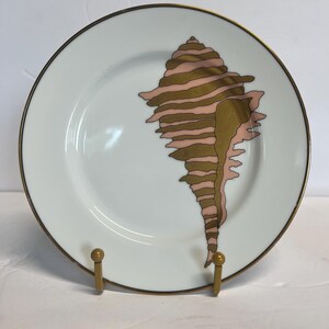 May include: White ceramic plate with a gold rim and a large seashell design in shades of pink and gold. The plate is displayed on a gold-colored stand, showcasing its decorative seashell motif.