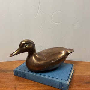 Op de afbeelding: Een bronzen eendenbeeldje met een glad, reflecterend oppervlak. De eend staat op een vintage blauw boek. Het beeldje heeft een lange snavel en een gedetailleerd oog, een klassiek decoratief stuk.