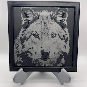 May include: A square black framed tile with a detailed grayscale image of a wolf's face. The wolf's fur is intricately depicted, with focused eyes and a prominent nose. The tile is displayed on a grey stand.