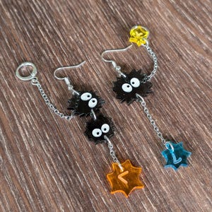 K&ouml;nnte beinhalten: Ohrringe mit schwarzen Cartoon-Charakteren mit wei&szlig;en Augen und einer Kette. Die Ohrringe haben gelbe, orangefarbene und blaue sternf&ouml;rmige Akzente. Auf einer Holzoberfl&auml;che.