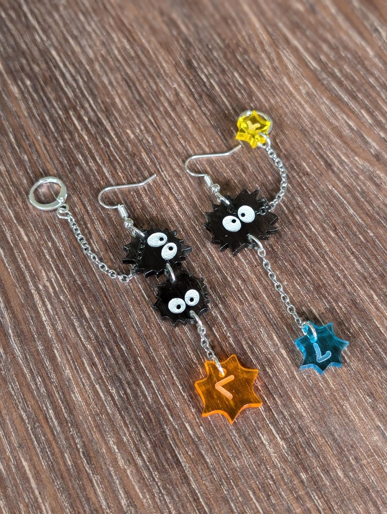 K&ouml;nnte beinhalten: Ohrringe mit schwarzen Cartoon-Charakteren mit wei&szlig;en Augen und einer Kette. Die Ohrringe haben gelbe, orangefarbene und blaue sternf&ouml;rmige Akzente. Auf einer Holzoberfl&auml;che.