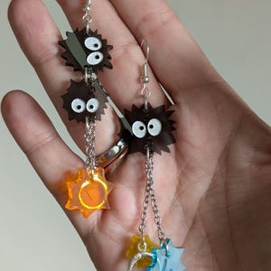 May include: A pair of dangle earrings featuring a whimsical design. The earrings have dark brown, spiky shapes with white eyes, connected by chains. Below, there are colorful star-shaped charms in orange, yellow, and blue. The earrings are held up by silver hooks.