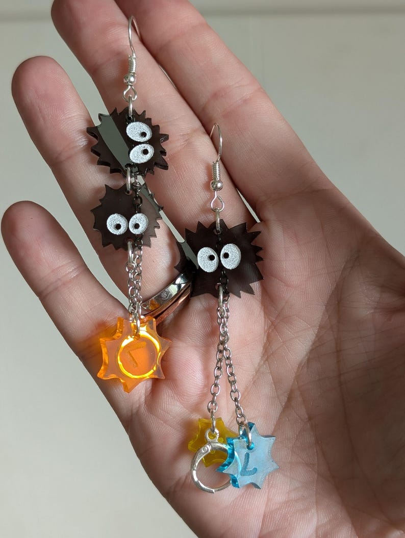 K&ouml;nnte beinhalten: Ohrringe zum H&auml;ngen mit einem verspielten Design. Die Ohrringe haben dunkelbraune, stachelige Formen mit wei&szlig;en Augen, die durch Ketten verbunden sind. Darunter befinden sich farbenfrohe sternf&ouml;rmige Anh&auml;nger in Orange, Gelb und Blau. Silberne Haken.