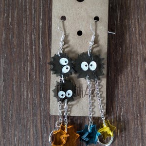 K&ouml;nnte beinhalten: Ohrringe mit schwarzen, stacheligen Cartoon-Charakteren mit wei&szlig;en Augen. Jeder Ohrring hat eine Kette mit sternf&ouml;rmigen Anh&auml;ngern in Orange, Blau und Gelb. Die Ohrringe h&auml;ngen an silberfarbenen Haken.