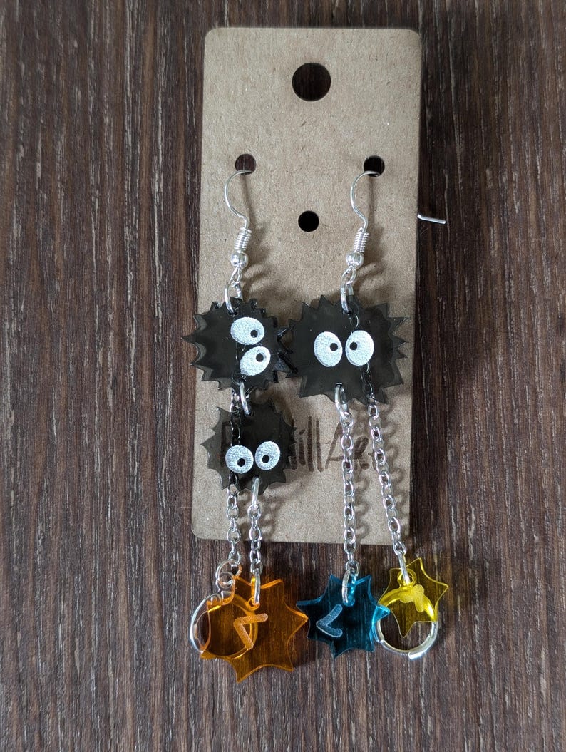 K&ouml;nnte beinhalten: Ohrringe mit schwarzen, stacheligen Cartoon-Charakteren mit wei&szlig;en Augen. Jeder Ohrring hat eine Kette mit sternf&ouml;rmigen Anh&auml;ngern in Orange, Blau und Gelb. Die Ohrringe h&auml;ngen an silberfarbenen Haken.