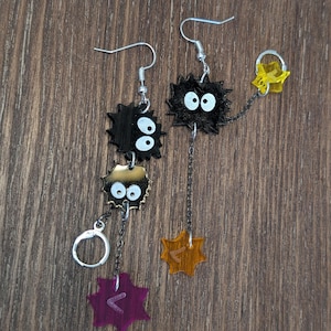 K&ouml;nnte beinhalten: Ein Paar einzigartige H&auml;ngeohrringe mit schwarzen, stacheligen, cartoonartigen Figuren mit wei&szlig;en Augen. Jeder Ohrring hat eine Kette mit einem Anh&auml;nger, einem lila und einem orangefarbenen, mit eingravierten Symbolen. Die Ohrringe h&auml;ngen an silberfarbenen Haken.