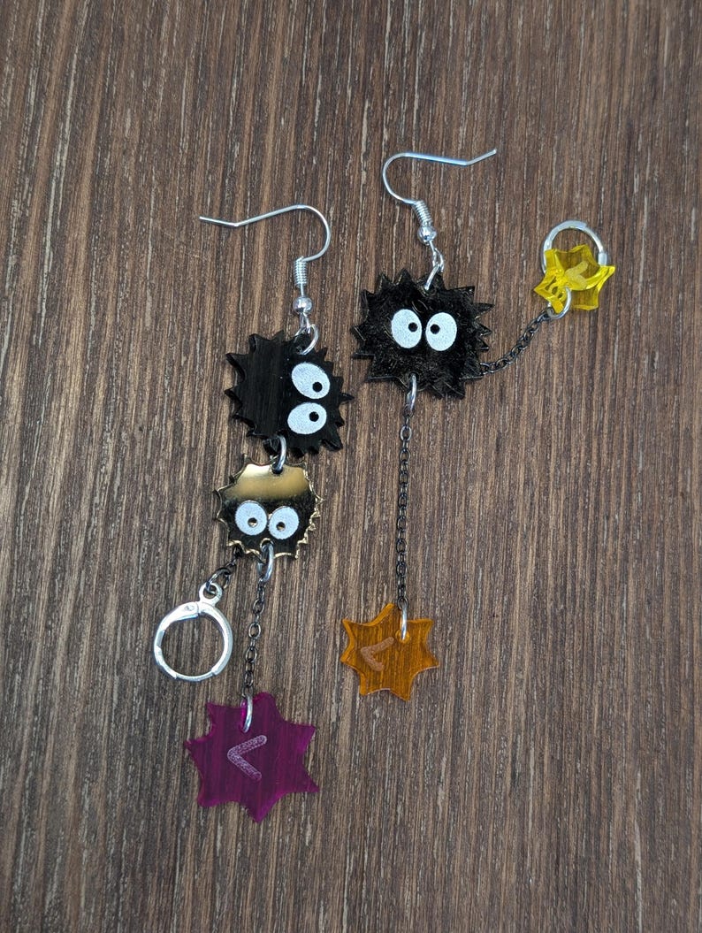 K&ouml;nnte beinhalten: Ein Paar einzigartige H&auml;ngeohrringe mit schwarzen, stacheligen, cartoonartigen Figuren mit wei&szlig;en Augen. Jeder Ohrring hat eine Kette mit einem Anh&auml;nger, einem lila und einem orangefarbenen, mit eingravierten Symbolen. Die Ohrringe h&auml;ngen an silberfarbenen Haken.