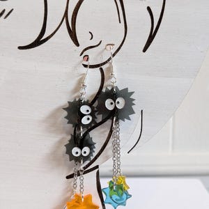 K&ouml;nnte beinhalten: Ohrringe zum Aufh&auml;ngen mit schwarzen, stacheligen Cartoon-Charakteren mit wei&szlig;en Augen und silbernen Kettenakzenten. Die Ohrringe haben orangefarbene und blaue Sternschnuppen-Anh&auml;nger. Auf wei&szlig;em Hintergrund.