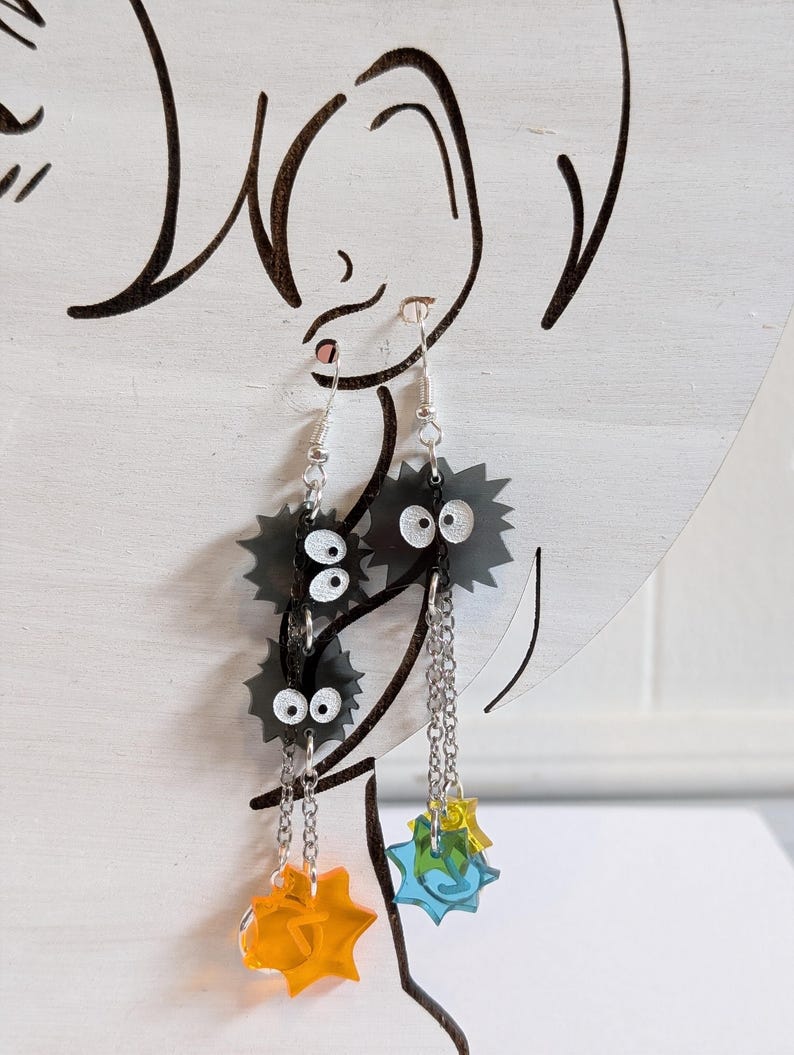 K&ouml;nnte beinhalten: Ohrringe zum Aufh&auml;ngen mit schwarzen, stacheligen Cartoon-Charakteren mit wei&szlig;en Augen und silbernen Kettenakzenten. Die Ohrringe haben orangefarbene und blaue Sternschnuppen-Anh&auml;nger. Auf wei&szlig;em Hintergrund.