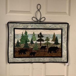 May include: A quilted wall hanging featuring a woodland scene with black bears and green trees against a blue and white background. The bears are walking through the forest, and the trees are tall and lush.