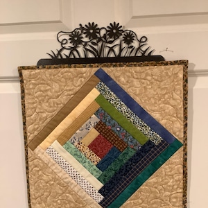 May include: A quilted wall hanging with a central diamond pattern composed of various fabric scraps in shades of blue, green, red, and beige. The quilt is framed by a patterned border and topped with a decorative metal floral accent.