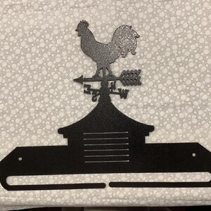 May include: Black metal rooster weather vane with a farmhouse style design. The vane features a rooster with a raised tail and a compass rose with the directions N, S, E, and W. The vane is mounted on a black metal base with a shelf.