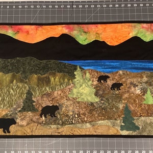 May include: A quilted wall hanging depicting a mountain landscape with a lake and a sunset sky. The scene features five black bears in silhouette, with trees and foliage in shades of green, brown, and orange.