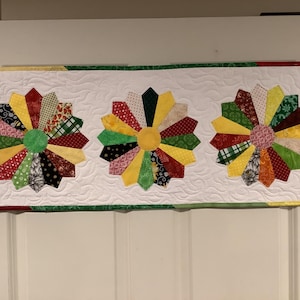 May include: A white quilted table runner with three colorful patchwork pinwheel designs in red, yellow, green, and black.