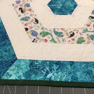 May include: A quilted, hexagonal table runner featuring a mermaid pattern on a white background. The border is a teal marble design with a dark green trim. The mermaid fabric has floral accents.