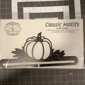 May include: Black metal craft holder with a pumpkin design. The holder is hand-crafted and has a bar for holding craft supplies. The text "Classic Motifs Craft Holder" is printed on the holder.