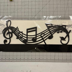 May include: Black metal music-themed wall decor featuring a treble clef, musical notes, and a bass clef. The design includes the text "Classic Motifs" and "Hand Crafted with Love". The decor is packaged in a clear plastic sleeve.