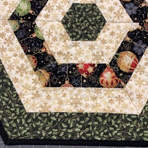 Può includere: Un runner da tavolo esagonale con sfondo nero che presenta un motivo di ornamenti dorati e foglie di agrifoglio. Il runner ha un bordo bianco con fiocchi di neve dorati e un bordo verde con un motivo di foglie di agrifoglio.