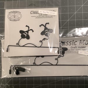 May include: Black metal craft holders with bee-shaped accents. The holders are designed for cross-stitch, quilting, and needlepoint projects. The image shows two holders, one labeled "5" Busy Bee Yellips" and the other "12" Busy Bee Yellips".