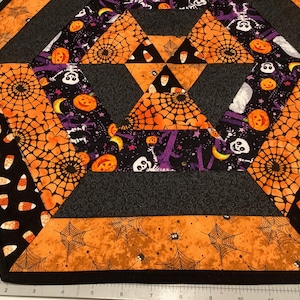 May include: Hexagon-shaped Halloween-themed table runner. The runner features a patchwork design with orange spiderwebs, candy corn, pumpkins, and skulls on a black and gray background. The edges are finished with a black border.