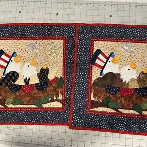 May include: Two patriotic eagle-themed quilted placemats. Each features a central design of eagles in a nest, wearing Uncle Sam hats, with a dotted background and a red, white, and blue color scheme. The borders are navy blue with white polka dots.