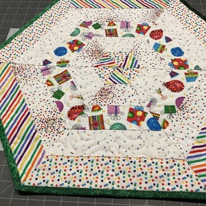 May include: A hexagonal table runner with a white background and a colorful confetti pattern. The runner has a green border and is made of fabric.