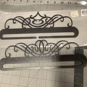 May include: Two decorative metal wall art pieces. The pieces are dark gray with an ornate, swirling design at the top and a horizontal bar with a curved opening. One piece is in a clear plastic bag. The pieces are on a gray cutting mat.