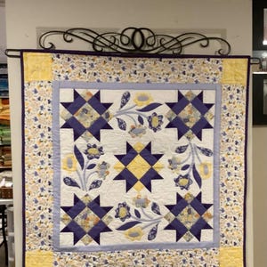 May include: A quilt with a white background and a pattern of purple stars and yellow flowers. The quilt is framed with a yellow and white floral border.