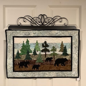 May include: A quilted wall hanging featuring a forest scene with evergreen trees and bears. The artwork has a black border and is mounted on a white door. A decorative metal scroll is above the artwork.