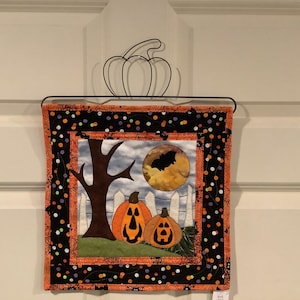 May include: Halloween-themed wall hanging featuring a spooky scene with two carved pumpkins, a tree, a white picket fence, and a full moon with a bat silhouette. The border is black with orange polka dots and spiderweb accents.