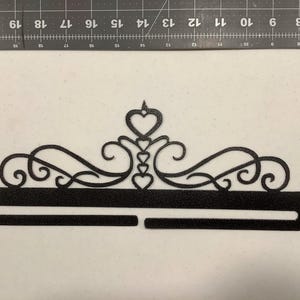 May include: Black metal medal display with a decorative heart design. The display has two horizontal bars for hanging medals.