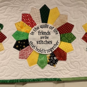 May include: A white quilt with a green border. The quilt has a large, colorful, star-shaped design in the center. The center of the star has a white circle with the text "In the quilt of life friends are the stitches that hold it together".