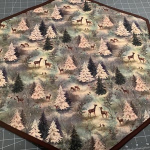 May include: Hexagon-shaped table runner with a winter forest print. The fabric features a repeating pattern of stylized pine trees, deer, and snowflakes on a blue and green background.