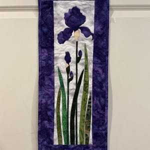 May include: A quilted wall hanging featuring a purple iris flower with green leaves and buds. The iris is centered on a white background, framed by a purple border. The fabric has a textured appearance.