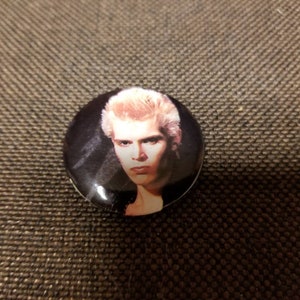 May include: A round button with a black background and a white border. The button features a colour photograph of a man with blonde hair and a serious expression.