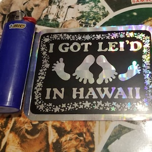 May include: A silver holographic sticker with a black background and white text that reads "I GOT LEI'D IN HAWAII". The sticker features three white footprints.