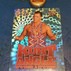 Puede incluir: Una tarjeta de intercambio holográfica con un luchador en un singlet de lucha rojo, blanco y azul. El luchador tiene los brazos extendidos hacia los lados. La tarjeta tiene el texto "Kurt Angle" en letras rojas grandes y "It's True It's True" en letras más pequeñas debajo. La tarjeta también tiene un pequeño logotipo "WF" en la esquina superior derecha.