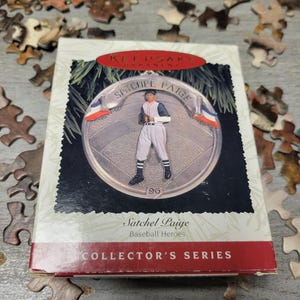 Könnte beinhalten: Eine "Keepsake Ornament"-Box mit einem Baseball-Ornament. Das Ornament zeigt Satchel Paige in einer Baseball-Uniform. Auf der Box steht "Collector's Series" und ist von Puzzleteilen umgeben.