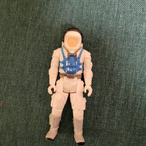 May include: A small, white astronaut action figure with a blue harness and helmet. The figure has a tan face and is wearing a white spacesuit with grey boots. The figure is standing upright.