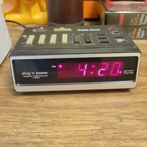 May include: A vintage Radio Shack plug 'n power remote controller timer with a digital display showing 4:20 PM. The black and silver device has several buttons and is on a wooden surface.
