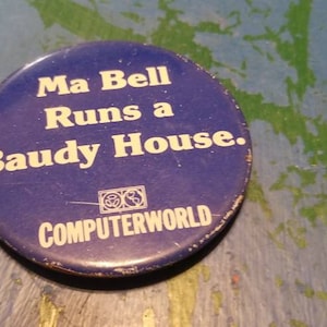 May include: A circular blue badge with white text: "Ma Bell Runs a Baudy House." The word "COMPUTERWORLD" and a graphic are below. The badge has a worn appearance, suggesting it is vintage.