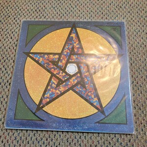 May include: A vinyl record album cover featuring a large, colorful star design. The star is filled with blue, orange, and brown shapes, set against a gold circle and a blue border. The album is in a protective sleeve.