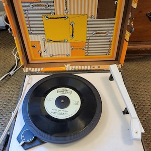 May include: A portable record player with a brown exterior and a lid featuring a yellow, orange, and white patchwork pattern. The turntable holds a black vinyl record labeled Sesame Street Name The Animal. It includes a white tone arm and controls.