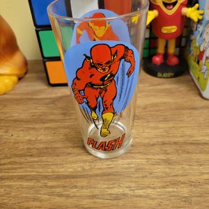 May include: A clear glass featuring a graphic of the Flash in a running pose, with a red and yellow costume on a blue background. The word "FLASH" is printed at the bottom of the glass. The glass is a vintage collectible.