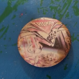 May include: A round button with a picture of the band Triumph. The button has a pink, white, and blue color scheme. The band's name is printed in white letters on a pink background.