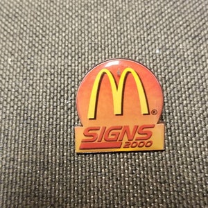 May include: A yellow and red enamel pin with the McDonald's golden arches logo and the text "SIGNS 2000".