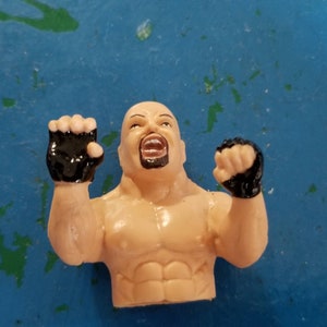Könnte beinhalten: Kleine Plastikfigur eines muskulösen, glatzköpfigen Mannes mit Ziegenbart, offenem Mund und erhobenen Fäusten. Die Figur hat schwarze Handschuhe und einen gebräunten Hautton. Der Hintergrund ist ein gesprenkeltes Blau.