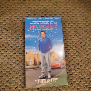 May include: A VHS tape for the movie "Mr. Destiny" featuring James Belushi and Michael Caine. The cover has a blue background with the title in large, colorful letters and a picture of a man. The phrase "Thumbs Up!" is at the bottom.