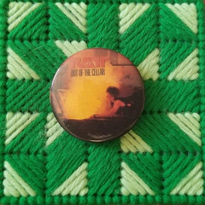 May include: A round button with a green, white, and yellow background. The button features a picture of a band called "Ram" with the text "Out of the Cellar".